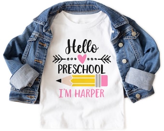 Puede incluir: Camiseta blanca con el texto "Hello Preschool" y un dibujo de lápiz. El nombre "I'm Harper" está impreso debajo del lápiz. Una chaqueta vaquera está sobre la camiseta.