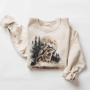 May include: A beige sweatshirt featuring a graphic print of two wolves howling at the moon in a woodland scene.