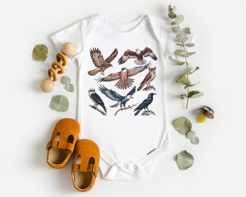 K&ouml;nnte beinhalten: Wei&szlig;er Baby-Body mit einer farbenfrohen Illustration verschiedener Greifv&ouml;gel, darunter Falken, Adler und eine Eule. Das Design zeigt die V&ouml;gel im Flug und auf &Auml;sten sitzend. Der Body ist von Babyschuhen und einem Holzspielzeug umgeben.