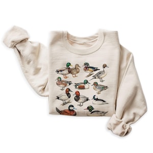 Könnte beinhalten: Cremefarbenes Sweatshirt mit einer detaillierten Illustration verschiedener Entenarten. Die Enten sind in einem realistischen Stil mit leuchtenden Farben dargestellt, darunter Grün, Braun und Weiß. Das Sweatshirt hat einen klassischen Rundhalsausschnitt und lange Ärmel.