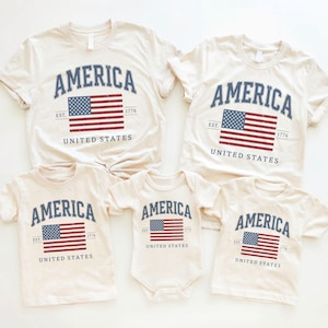 Puede incluir: Camisetas y un body de color crema con la palabra "AMERICA" en azul marino arqueada. Debajo, un rectángulo azul marino con la bandera estadounidense roja y blanca y el texto "EST. 1776 UNITED STATES".