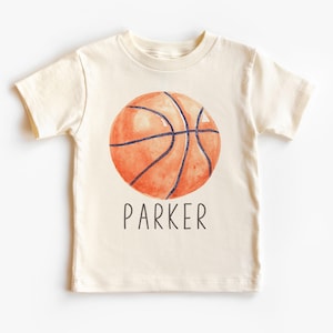 May include: Cream-colored t-shirt featuring a watercolor basketball design in shades of orange and brown, with black lines. The name "PARKER" is printed in black below the basketball. A short-sleeved shirt.