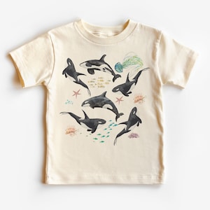May include: A cream-colored t-shirt with a graphic print of black and white orcas swimming in a blue ocean with colorful fish, starfish, and jellyfish.