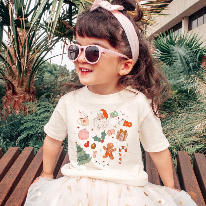 Puede incluir: Una ni&ntilde;a lleva una camiseta color crema con un dise&ntilde;o navide&ntilde;o. El dise&ntilde;o incluye a Pap&aacute; Noel, un hombre de jengibre y la palabra "HO". La ni&ntilde;a tambi&eacute;n lleva gafas de sol rosas y una diadema rosa.