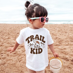 Peut inclure: T-shirt blanc avec l'inscription "TRAIL KID" en marron, repr&eacute;sentant une ch&egrave;vre de dessin anim&eacute; avec un sac &agrave; dos et un motif de montagne. L'enfant porte des lunettes de soleil roses et se tient sur une plage de sable.