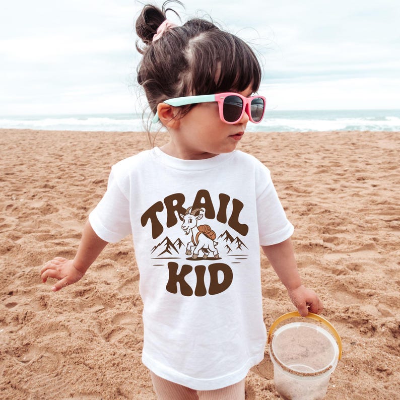 Peut inclure: T-shirt blanc avec l'inscription "TRAIL KID" en marron, repr&eacute;sentant une ch&egrave;vre de dessin anim&eacute; avec un sac &agrave; dos et un motif de montagne. L'enfant porte des lunettes de soleil roses et se tient sur une plage de sable.