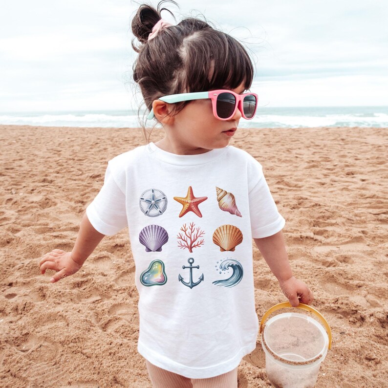 May include: A white t-shirt featuring colorful watercolor illustrations of seashells, a starfish, an anchor, and a wave. The shirt is worn by a young child on a sandy beach, with the ocean visible in the background.
