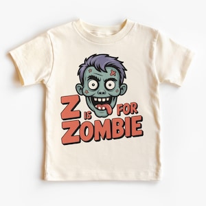 Puede incluir: Camiseta crema para niños pequeños con un dibujo de una cara de zombi con pelo morado y el texto "Z is for Zombie" en naranja y rojo. El zombi tiene piel verde, una lengua que sobresale y dientes visibles.