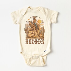 May include: A beige baby onesie with a vintage illustration of a cowboy riding a horse in a desert landscape. The onesie has the name "HUDSON" printed in brown text.