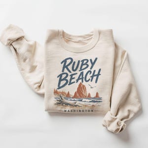 May include: A cream-colored crewneck sweatshirt featuring a graphic print of Ruby Beach, Washington. The design includes the text Ruby Beach in dark blue, a coastal landscape with sea stacks, driftwood, and a bird in shades of blue and brown.