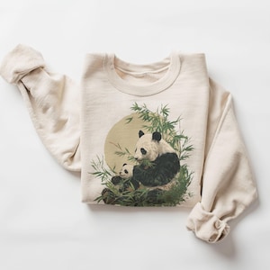 May include: Cream-colored sweatshirt featuring a panda bear and cub amidst bamboo. The graphic includes a yellow sun in the background. The long sleeves are rolled up. The sweatshirt is made of a soft material.