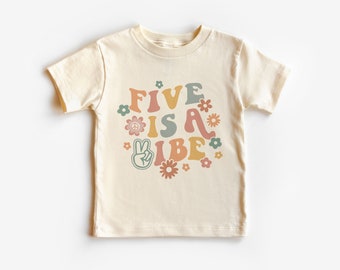 May include: Cream-colored t-shirt with the text "FIVE IS A VIBE" in retro-style lettering. The design includes flowers and a peace sign. The shirt is short-sleeved and suitable for children.