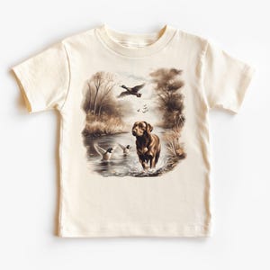 Pode incluir: Uma camiseta de cor creme com um labrador retriever marrom em pé em um riacho com patos voando acima. O design é uma pintura em aquarela.