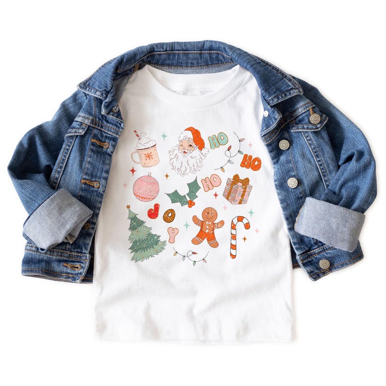 Puede incluir: Camiseta blanca con un dise&ntilde;o navide&ntilde;o que incluye a Pap&aacute; Noel, un &aacute;rbol de Navidad, un hombre de jengibre y un bast&oacute;n de caramelo. La camiseta se combina con una chaqueta vaquera azul.