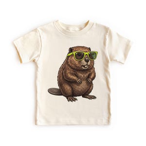 May include: Cream-colored t-shirt featuring a cartoon beaver wearing neon green sunglasses. The beaver is brown with detailed fur and a small, flat tail. The design is centered on the shirt, which has short sleeves.