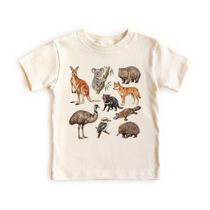 May include: Cream-colored t-shirt featuring illustrations of various Australian animals, including a kangaroo, koala, wombat, dingo, Tasmanian devil, platypus, emu, kookaburra, and echidna. The design is centered on the front of the shirt.
