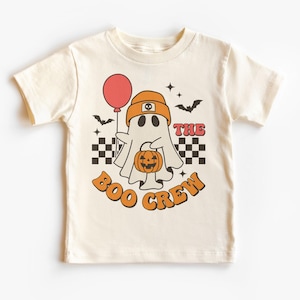 May include: Cream-colored t-shirt featuring a cartoon ghost wearing an orange beanie and holding a red balloon and a pumpkin. The text "THE BOO CREW" is in orange and red. Black and white checkered squares and bats complete the Halloween design.
