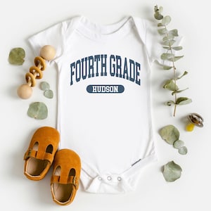 May include: White baby bodysuit with the text "FOURTH GRADE HUDSON" in navy blue. The image includes a pair of brown leather baby shoes, a wooden toy, and decorative greenery.