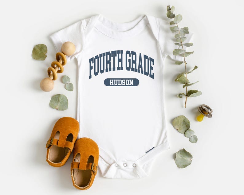 May include: White baby bodysuit with the text "FOURTH GRADE HUDSON" in navy blue. The image includes a pair of brown leather baby shoes, a wooden toy, and decorative greenery.