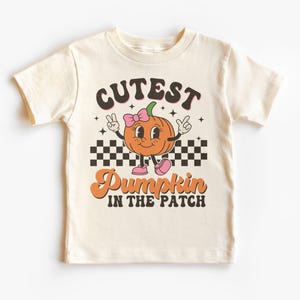 Puede incluir: Camiseta color crema con un gráfico retro. El diseño incluye el texto "Cutest Pumpkin in the Patch" sobre un personaje de calabaza naranja sonriente con un lazo y zapatos rosas, y un patrón de tablero de ajedrez.