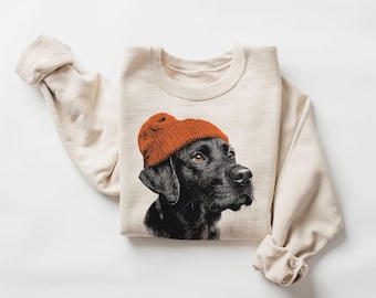 Può includere: Una felpa girocollo color crema con una stampa grafica dettagliata di un cane Labrador nero che indossa un berretto in maglia arancione brillante. Il cane è raffigurato di profilo, rivolto verso il lato destro del capo.