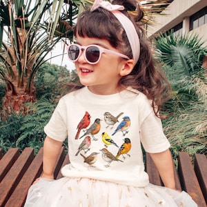 以下が含まれることがあります： さまざまな鳥のイラストが描かれたクリーム色のTシャツを着た幼い子供。子供はピンクのサングラスと、お揃いのヘアバンドも着用しています。子供は屋外の木製のベンチに座っています。