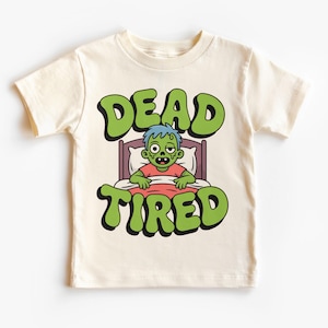 Puede incluir: Camiseta color crema con un gráfico verde y negro que dice "DEAD TIRED". El gráfico presenta un zombi de dibujos animados en la cama. El zombi tiene piel verde, pelo azul y lleva una camisa roja.