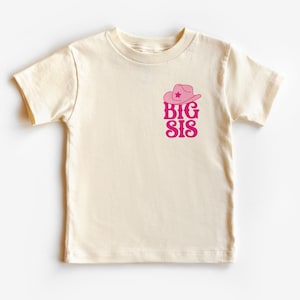 Puede incluir: Camiseta color crema con un gráfico de sombrero de vaquero rosa y las palabras "BIG SIS" en una fuente retro. El diseño está centrado en el pecho. La camiseta tiene mangas cortas y cuello redondo.