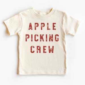 Könnte beinhalten: Cremefarbenes T-Shirt mit dem roten Aufdruck "APPLE PICKING CREW" in Blockbuchstaben. Der Text hat einen Used-Look. Das kurzärmelige Shirt scheint aus weicher Baumwolle zu sein.