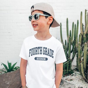 May include: A white t-shirt with the text "FOURTH GRADE HUDSON" in navy blue. The child is wearing a beige baseball cap and light green sunglasses. A cactus and other greenery are in the background.