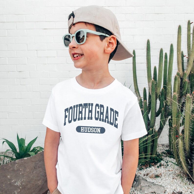 May include: A white t-shirt with the text "FOURTH GRADE HUDSON" in navy blue. The child is wearing a beige baseball cap and light green sunglasses. A cactus and other greenery are in the background.