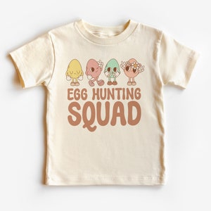 Puede incluir: Una camiseta de manga corta de color crema con un gráfico de cuatro huevos de dibujos animados que llevan diferentes expresiones. El texto "Egg Hunting Squad" está impreso en marrón debajo del gráfico.
