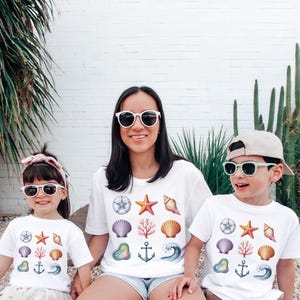 May include: Three people wearing white t-shirts with a colorful seashell design. The design includes a starfish, seashells, coral, and an anchor. All three are wearing sunglasses. The background is a white brick wall with green plants.