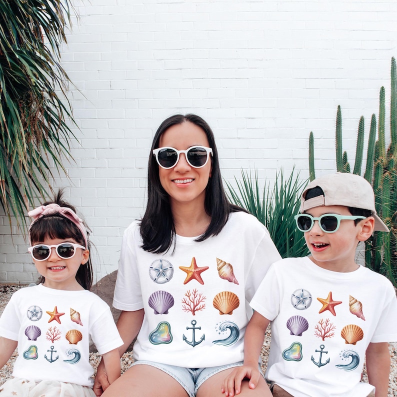 May include: Three people wearing white t-shirts with a colorful seashell design. The design includes a starfish, seashells, coral, and an anchor. All three are wearing sunglasses. The background is a white brick wall with green plants.