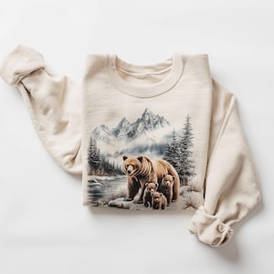 May include: A beige sweatshirt featuring a printed design of a brown bear and two cubs in a snowy mountain landscape.