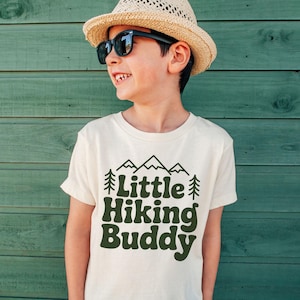 May include: A cream-colored t-shirt with the text "Little Hiking Buddy" in dark green, along with mountain and tree graphics. The child is wearing a straw hat and sunglasses.