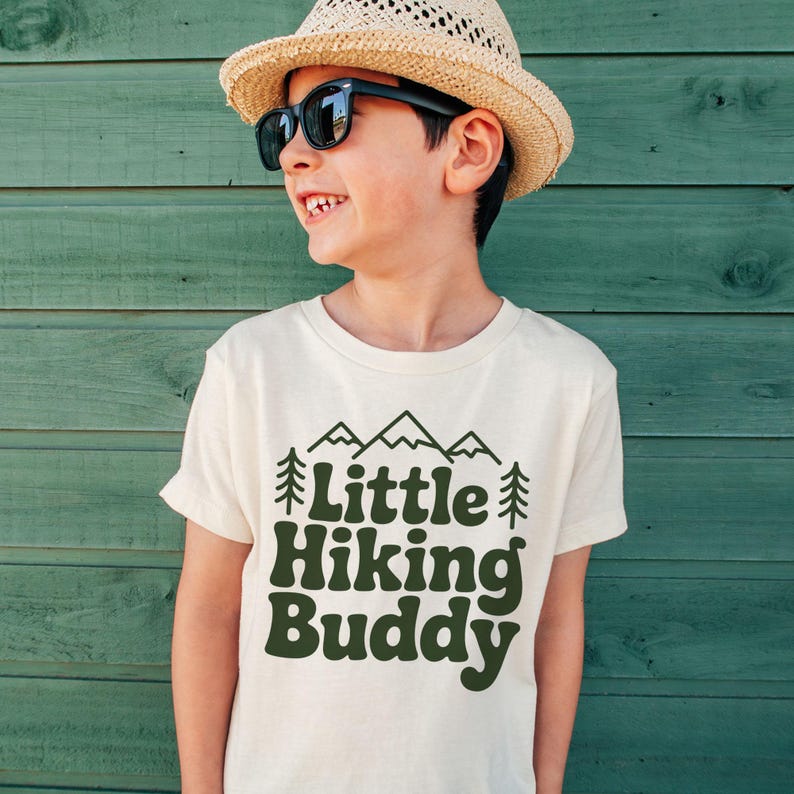 May include: A cream-colored t-shirt with the text "Little Hiking Buddy" in dark green, along with mountain and tree graphics. The child is wearing a straw hat and sunglasses.