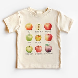 Peut inclure: Un t-shirt beige avec un graphique de neuf types de pommes différents. Chaque pomme est étiquetée avec son nom : Gala, Granny Smith, Honeycrisp, Crispin, Pink Pearl, Fuji, Ginger Gold, Red Delicious et Black Diamond.