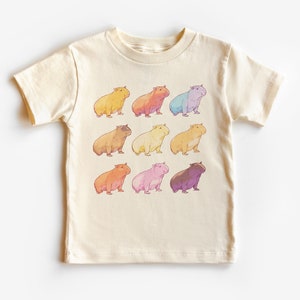 May include: A cream-colored t-shirt with a graphic of nine capybaras in different pastel colors. The capybaras are arranged in three rows of three.