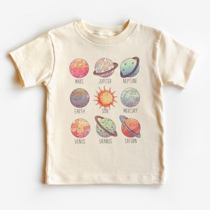 May include: A cream colored t-shirt with a graphic of the planets in our solar system. The planets are drawn in a colorful, cartoon style. The text "Mars", "Jupiter", "Neptune", "Earth", "Sun", "Mercury", "Venus", "Uranus", and "Saturn" are printed below each planet.