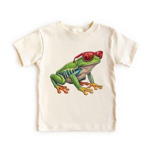May include: A cream-colored short-sleeve t-shirt featuring a graphic print of a green frog wearing red sunglasses. The frog has blue and yellow markings on its sides and orange-tipped toes, positioned centrally on the front of the garment.