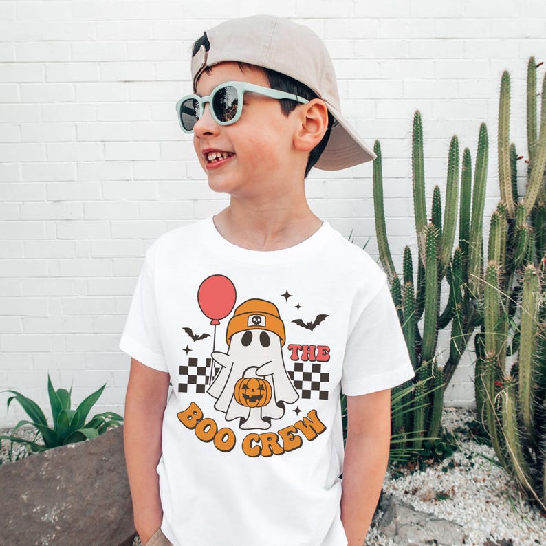 May include: White t-shirt with a Halloween-themed graphic. The design features a ghost wearing an orange hat and holding a red balloon and a pumpkin. The text "THE BOO CREW" is printed in orange.