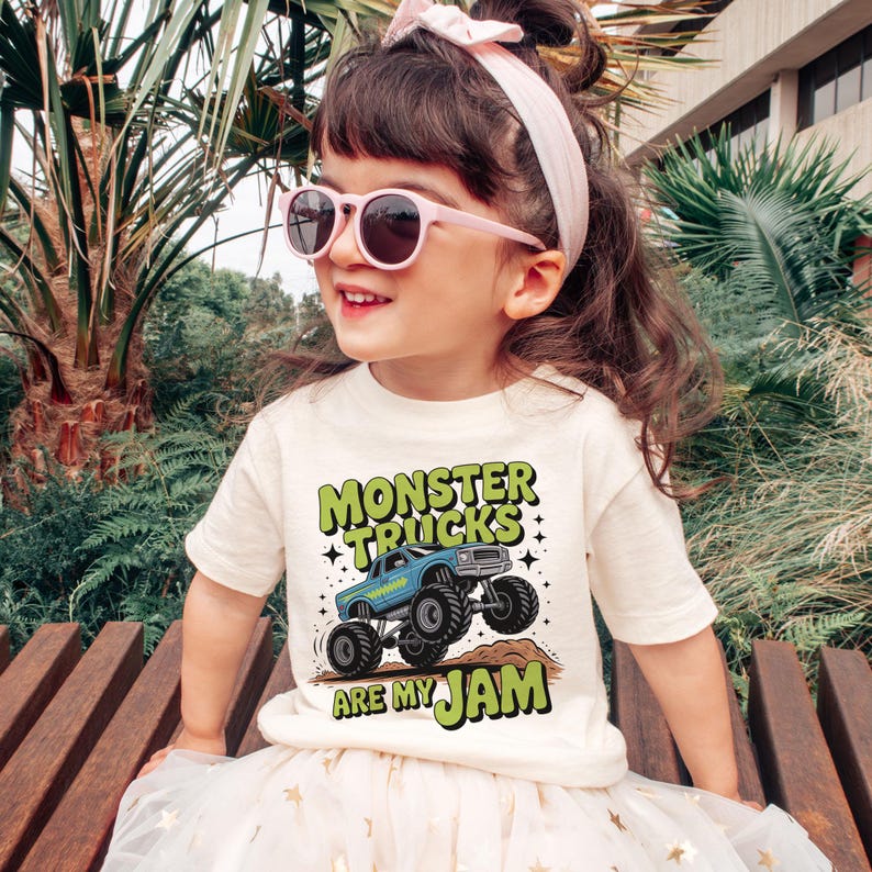 Puede incluir: Una camiseta color crema con el texto "MONSTER TRUCKS ARE MY JAM" en verde y azul. La camiseta presenta un gr&aacute;fico de un cami&oacute;n monstruo azul. La ni&ntilde;a lleva gafas de sol rosas y una diadema rosa.
