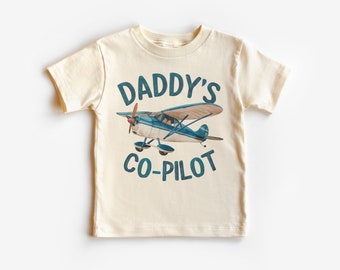 May include: Cream-colored short-sleeve t-shirt with the text "DADDY'S CO-PILOT" in teal. The shirt features a watercolor illustration of a blue and brown airplane. The design is centered on the front of the shirt.