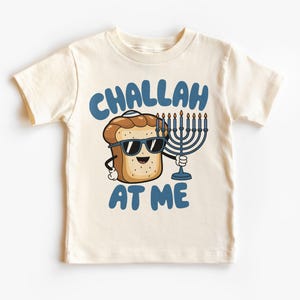 Puede incluir: Camiseta color crema con un personaje de pan challah de dibujos animados con gafas de sol y una menorá. El texto azul "CHALLAH AT ME" crea un diseño divertido y festivo.