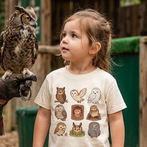 May include: A young child wearing a cream-colored t-shirt featuring nine different cartoon owl illustrations. A large owl with brown and white feathers is perched on a gloved hand. The background includes wooden structures and greenery.
