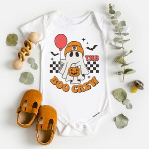May include: White baby onesie with a retro-style graphic of a ghost holding a pumpkin and a red balloon. The text "THE BOO CREW" is printed in orange. Includes a pair of brown baby shoes and wooden toys.