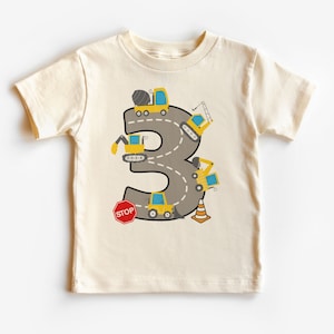 Peut inclure: Un t-shirt beige à manches courtes avec un graphique d'un numéro 3 fait d'une route grise avec des véhicules de chantier jaunes qui y circulent. Les véhicules comprennent un camion à béton, une grue, une pelle mécanique, un camion à benne basculante et un panneau d'arrêt.