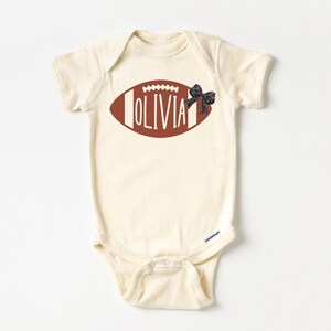 Puede incluir: Body de beb&eacute; color crema con un dise&ntilde;o de bal&oacute;n de f&uacute;tbol americano marr&oacute;n. El bal&oacute;n tiene costuras blancas y el nombre "OLIVIA" en blanco. Se adjunta un lazo negro. El body tiene mangas cortas y cierres a presi&oacute;n.