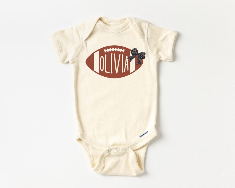 Puede incluir: Body de beb&eacute; color crema con un dise&ntilde;o de bal&oacute;n de f&uacute;tbol americano marr&oacute;n. El bal&oacute;n tiene costuras blancas y el nombre "OLIVIA" en blanco. Se adjunta un lazo negro. El body tiene mangas cortas y cierres a presi&oacute;n.